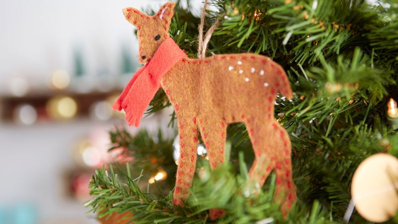 A hand-stitched deer wearing a red scarf ornament hanging on a Christmas tree.