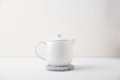 A white teapot on a finger knit grey trivet against a white background.