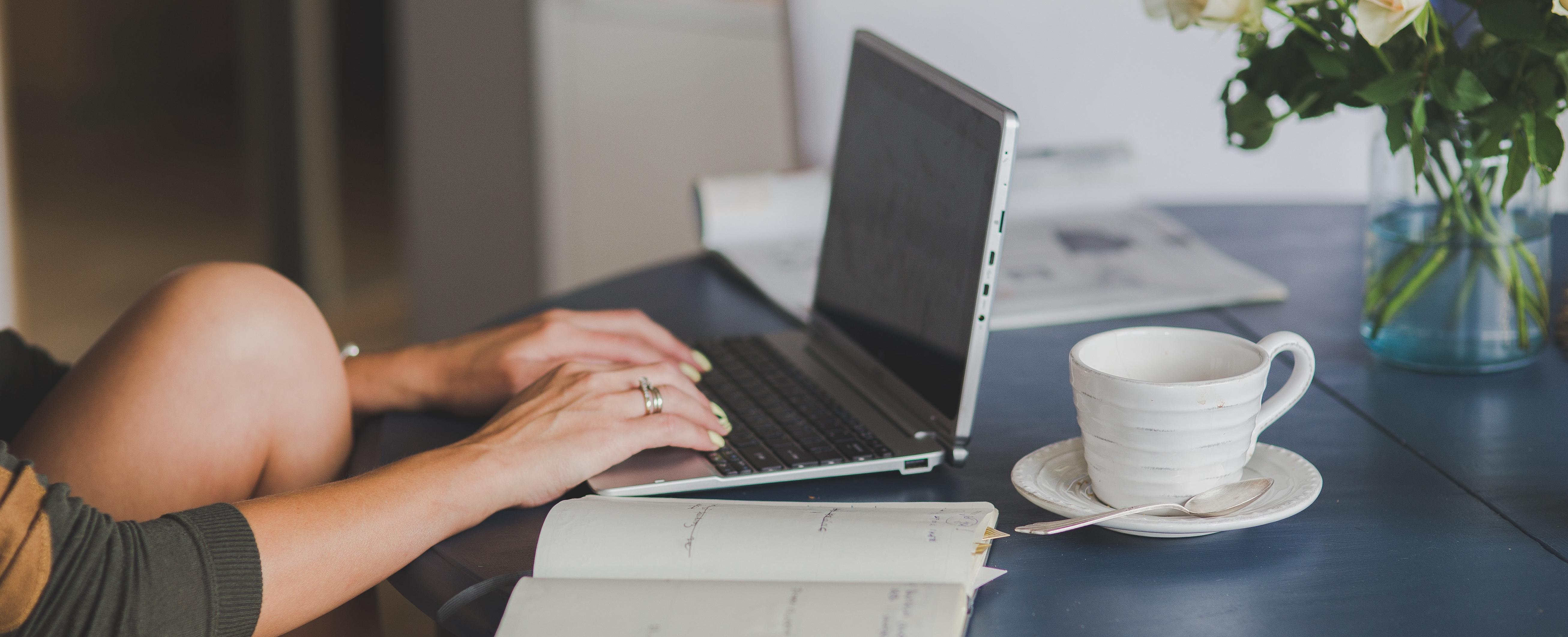 Person working on laptop at table with a notepad and mug